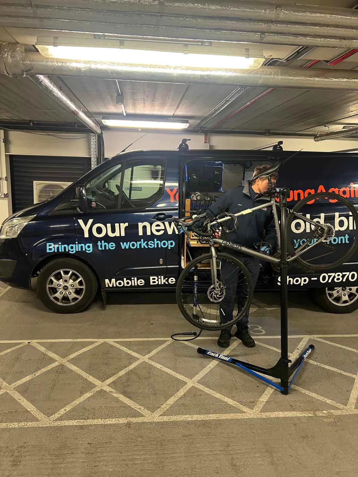 A bike on a portable repair stand beside the YoungAgain Cycles mobile workshop van, with the mechanic working behind it