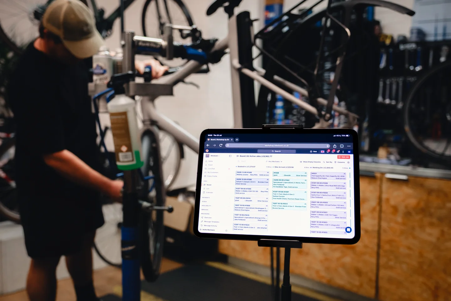 The Workshop job board open on a tablet in the Zelo Bikes workshop, with a mechanic at the repair stand behind it